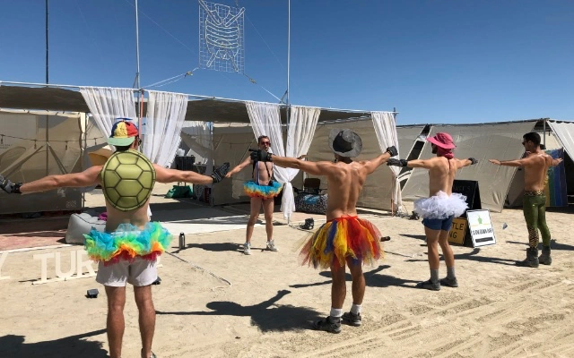 Young men in tutus doing aerobics in front of a dusty theme camp at Burning Man